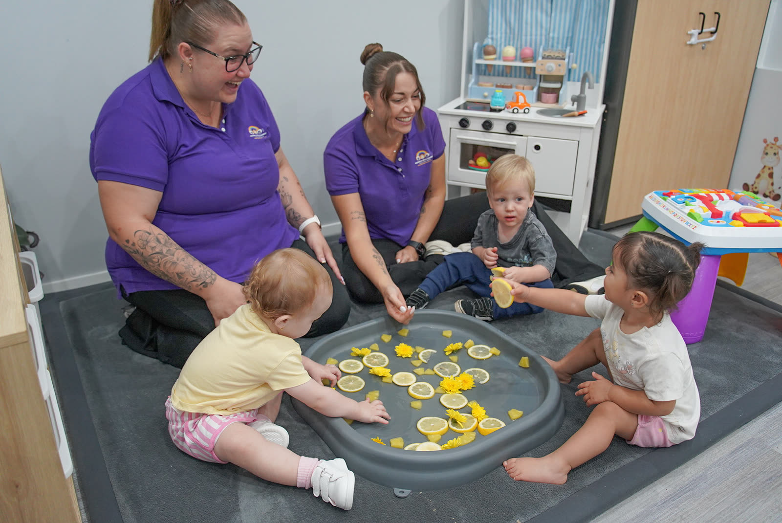 Two educators sitting on the floor with three babies exploring a tray of lemon slices and yellow flowers in water.