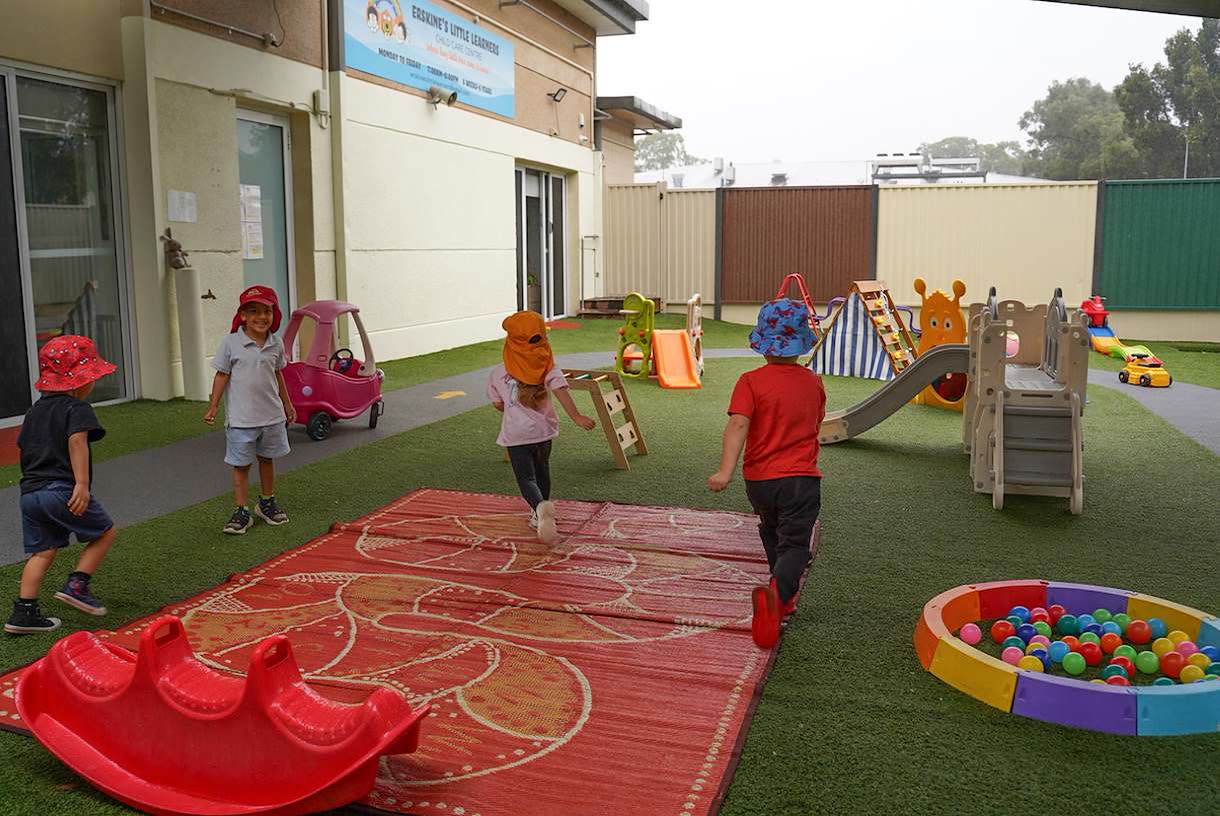 Four children in sun hats running and riding across the centre's grassed outdoor play area, past a slide and a ball pit.