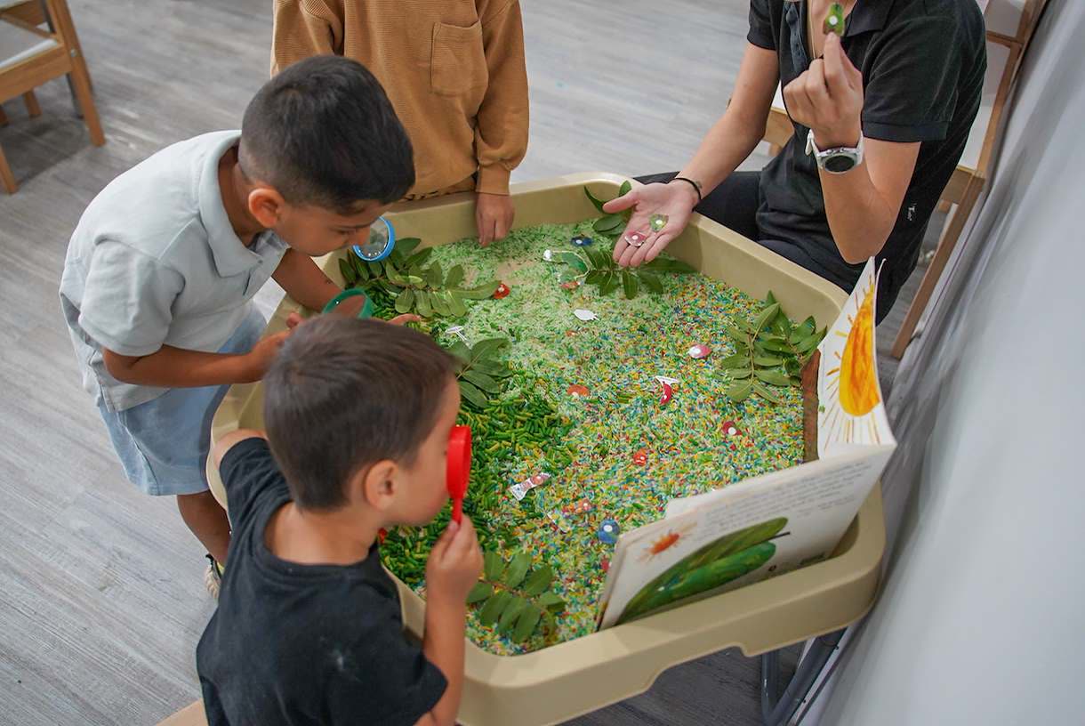 Children using magnifying glasses to look at leaves and coloured rice in a sensory tray, beside an open picture book.