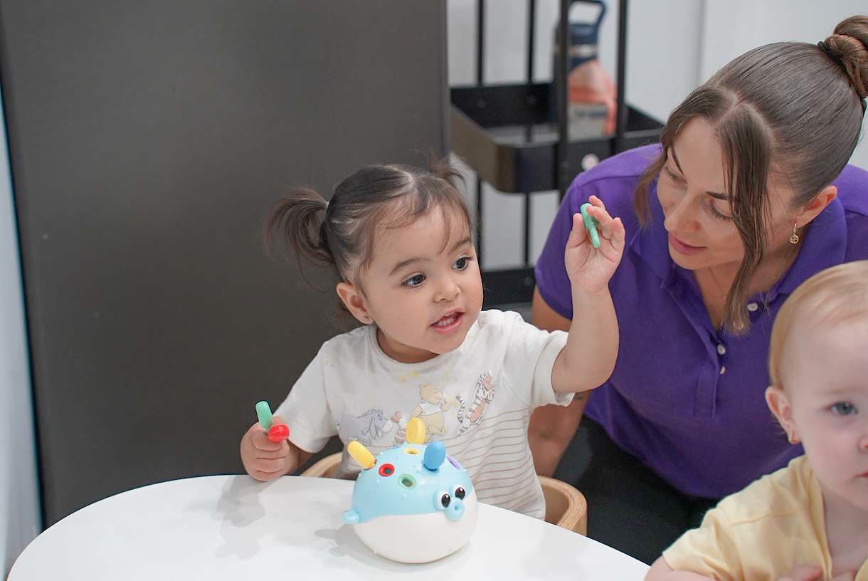 An educator leaning in beside a toddler who is posting coloured pegs into a toy at the table.