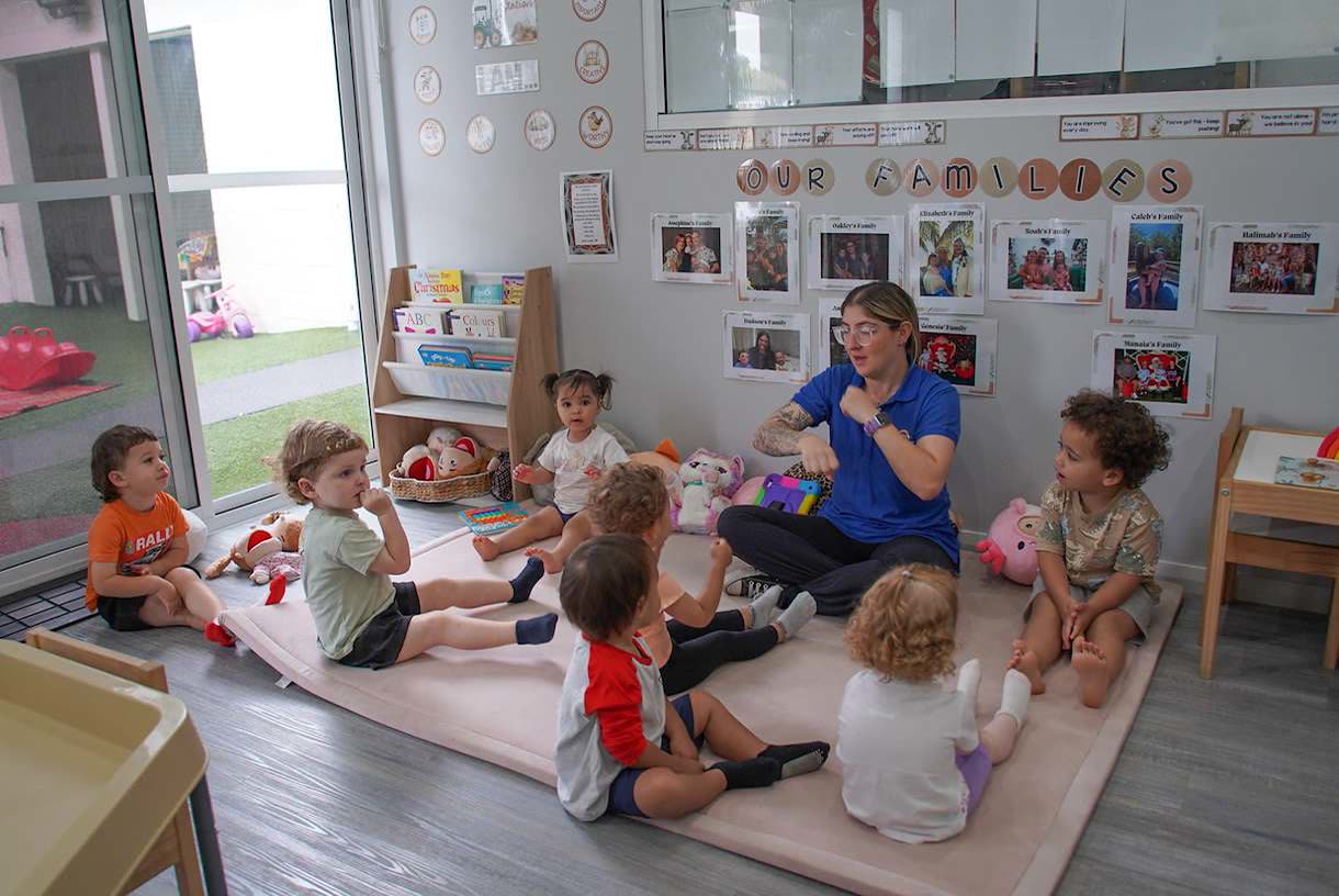 An educator signing to seven children sitting on a mat at group time, below a wall of family photographs.