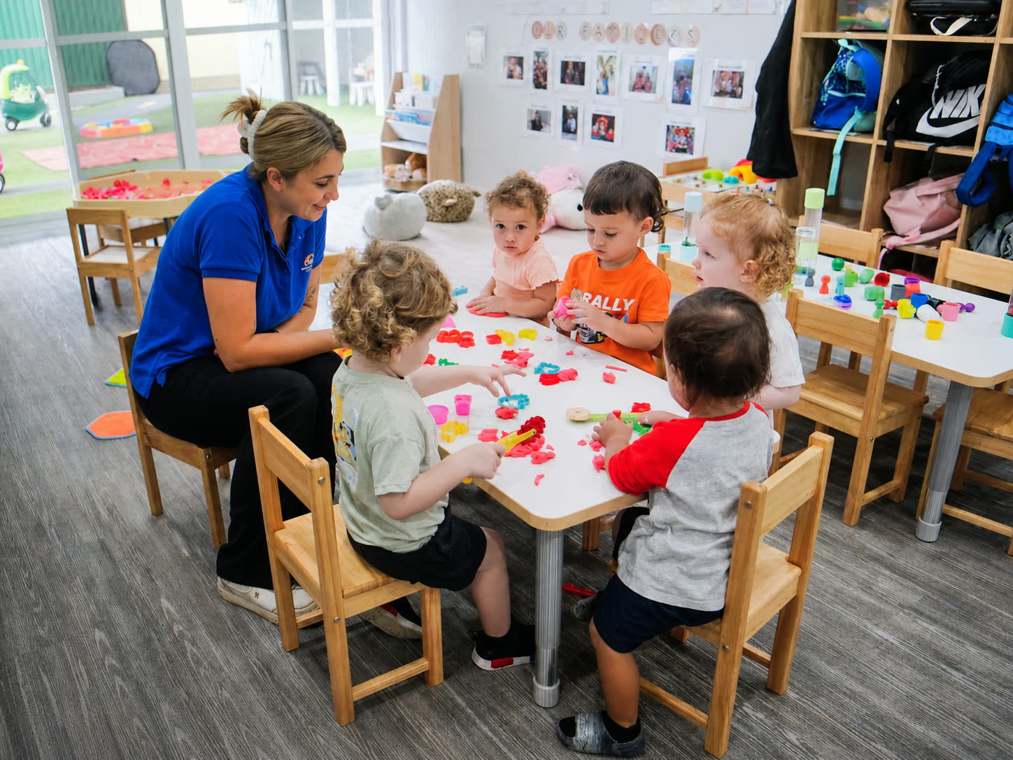 An educator at a low table with five toddlers rolling and cutting coloured play dough.